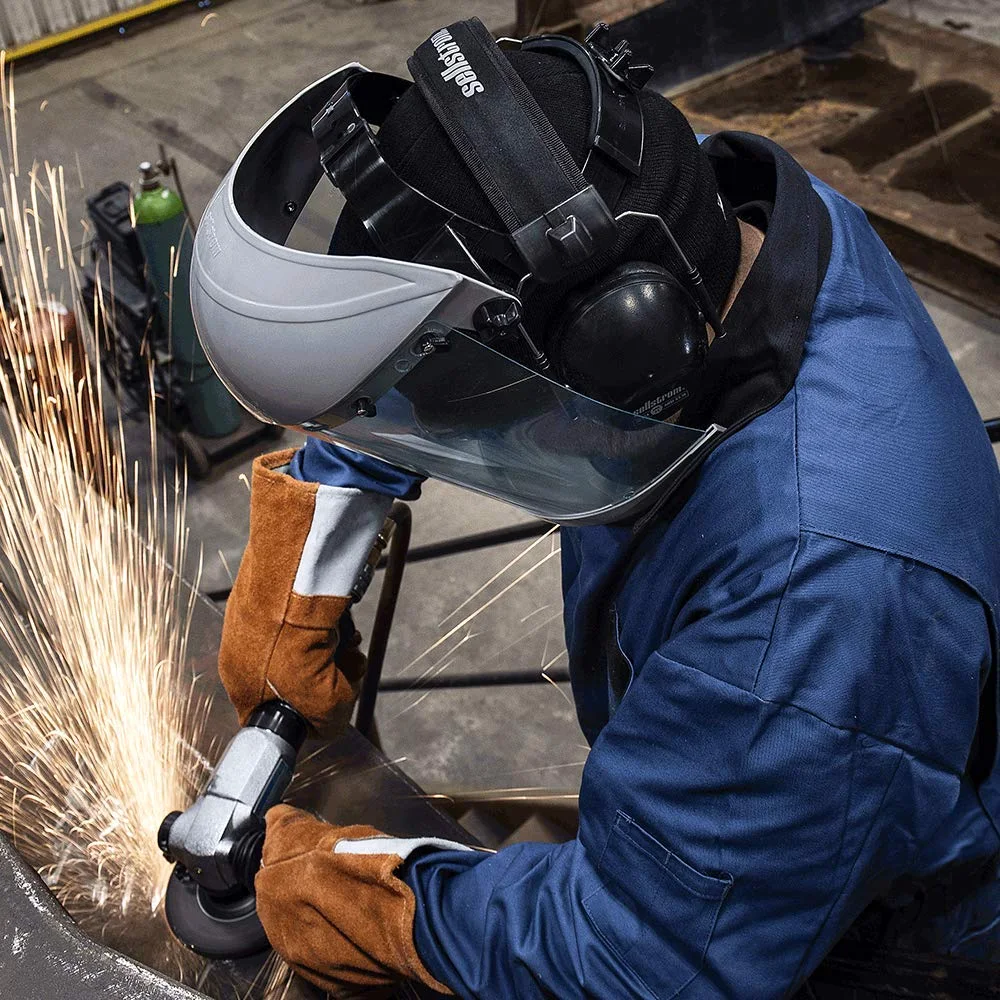Worker in a navy coverall grinding metal with an angle grinder, wearing a face shield with a gray crown, ear muffs and leather gloves as sparks fly