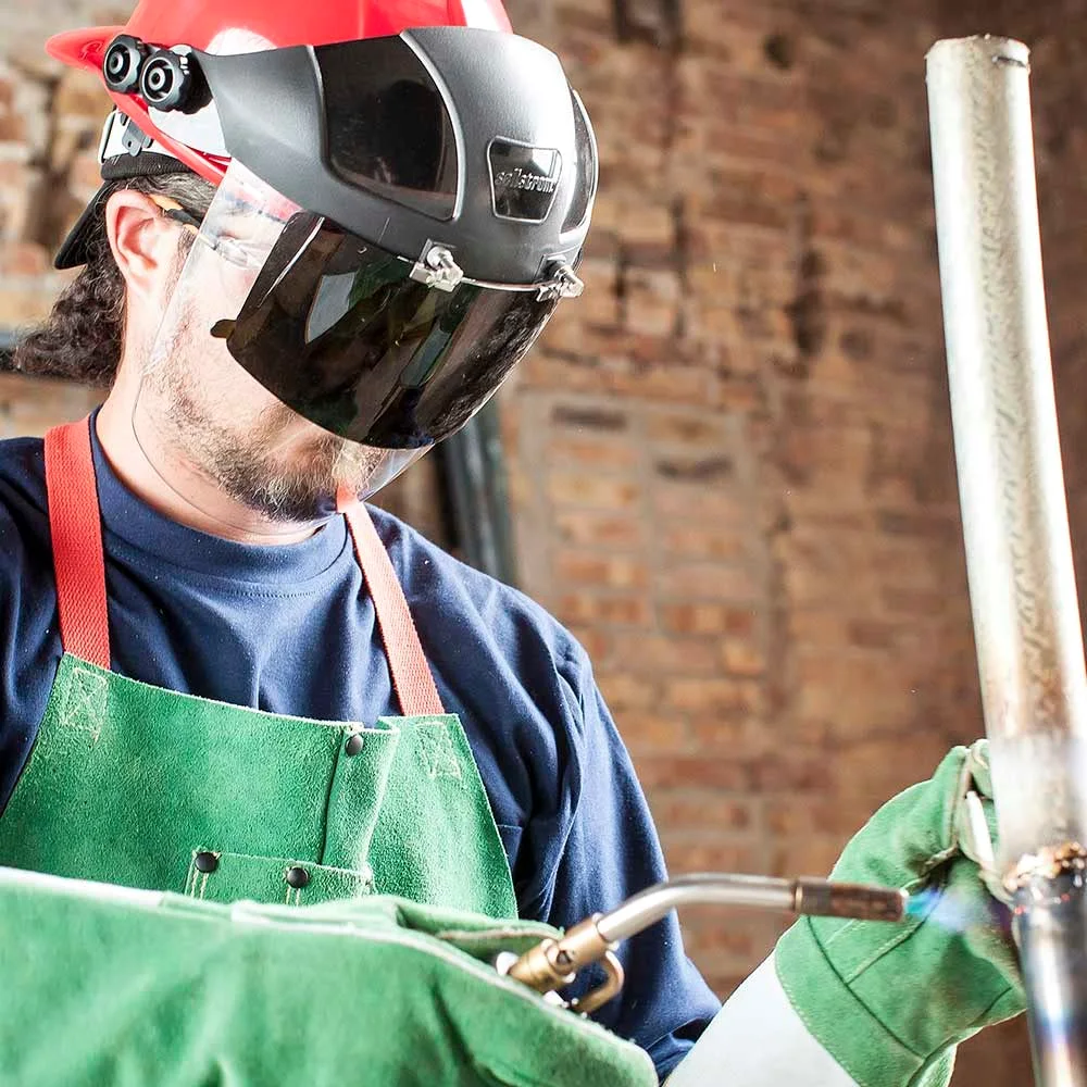 Worker in a red hard hat with a face shield and a dark tinted flip window, brazing a pipe with a torch, in a green leather apron and gloves