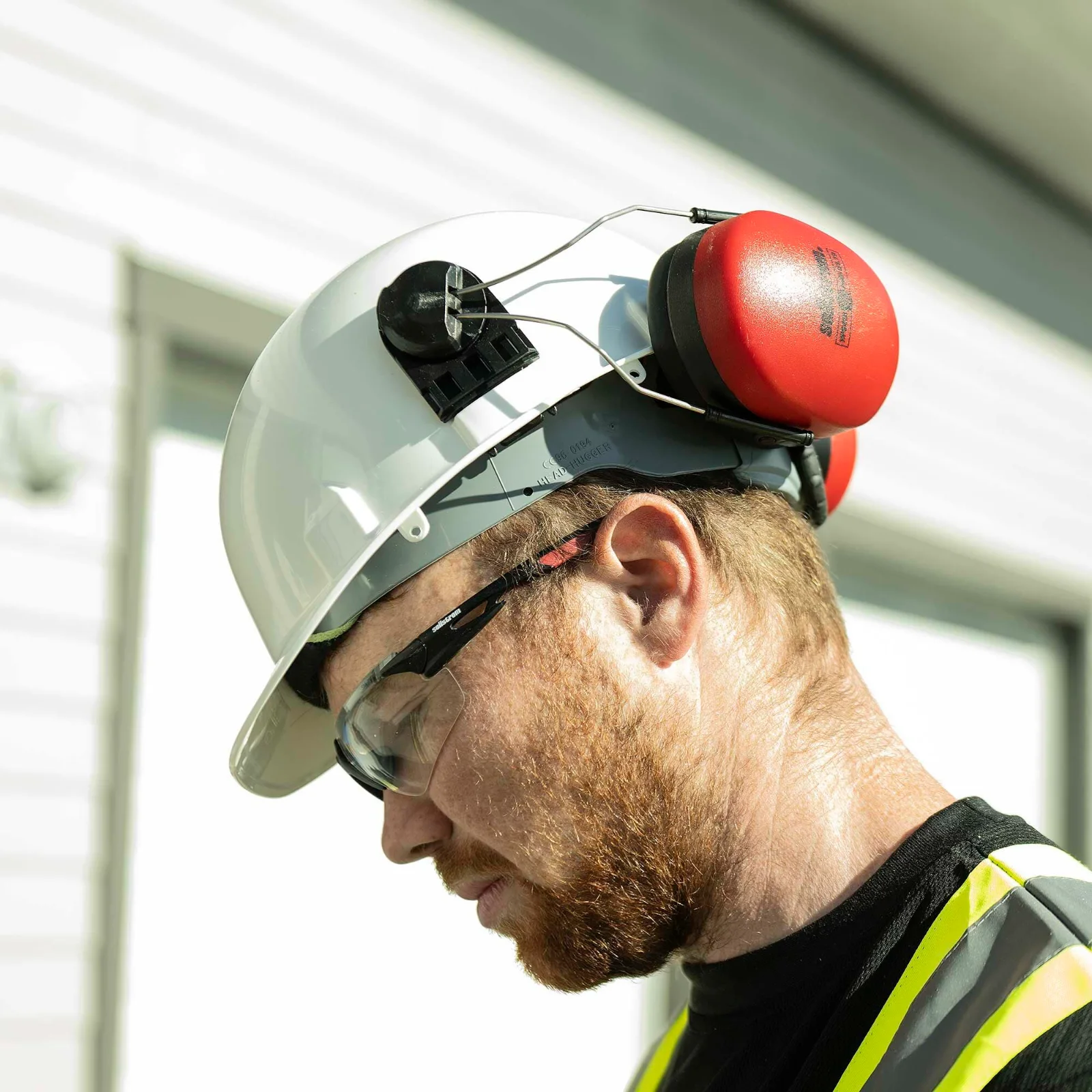 A man in a white hard hat and safety glasses with a red ear muff cup raised above his ear